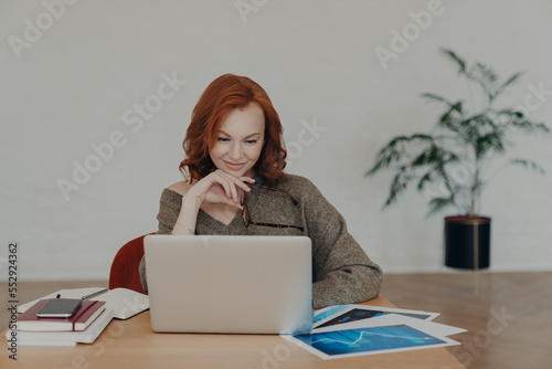 Busy woman freelancer holds spectacles for vision correction, poses at home desktop, has paperwork and works on laptop computer, concentrated at notebook screen. Technology and communication