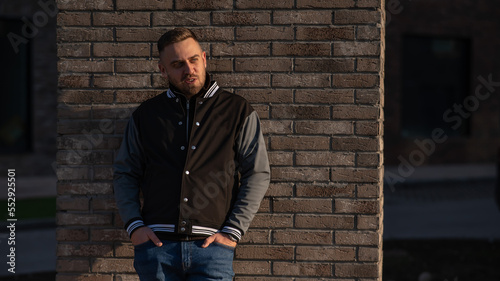 Caucasian bearded man in a bomber jacket leaned against a brick wall. 