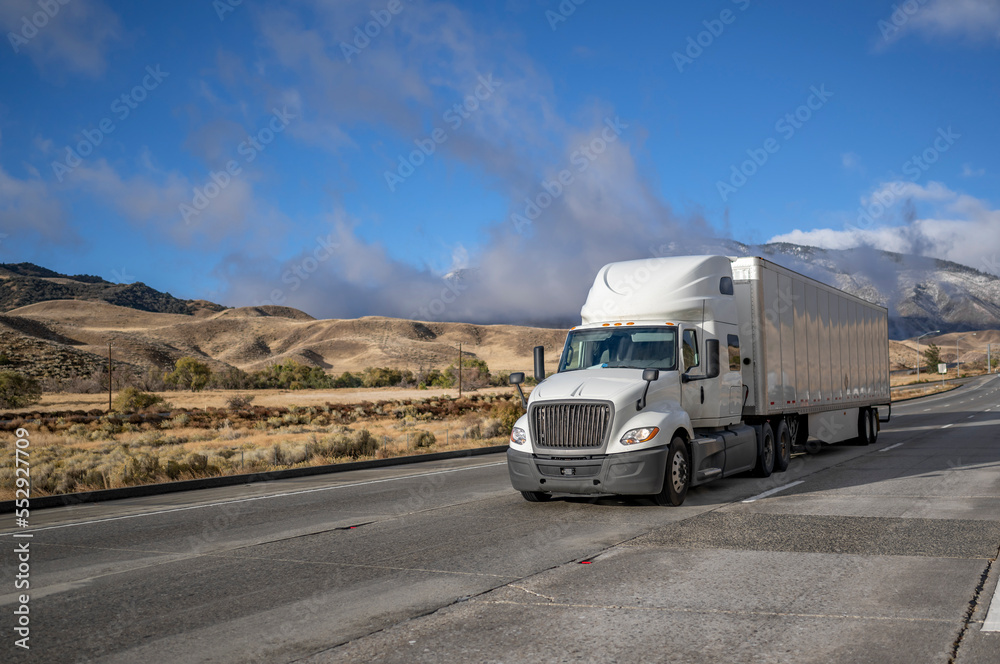 Bonnet industrial grade white big rig long hauler semi truck transporting cargo in dry van semi trailer running on the wide highway road with snow mountain and clouds on the background