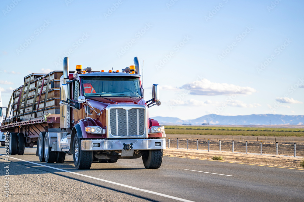 Burgundy day cab big rig semi truck transporting fastened commercial cargo on flat bed semi trailer driving on the straight highway road