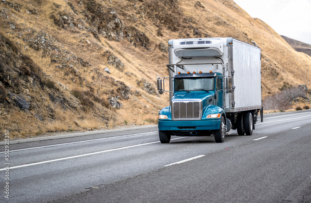 Obraz premium Compact day cab big rig semi truck with refrigerated box trailer transporting cargo driving on the wide multiline highway road with rocky hills on the sides
