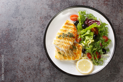 Fotografija Spicy grilled cod fish fillet with fresh vegetable salad and leaf lettuce mix and lemon close-up in a plate on the table