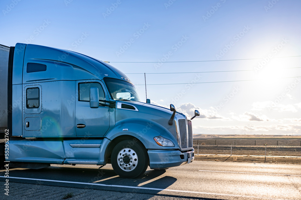 Powerful blue bonnet big rig semi truck tractor transporting cargo in dry van semi trailer running on the road at sunshine
