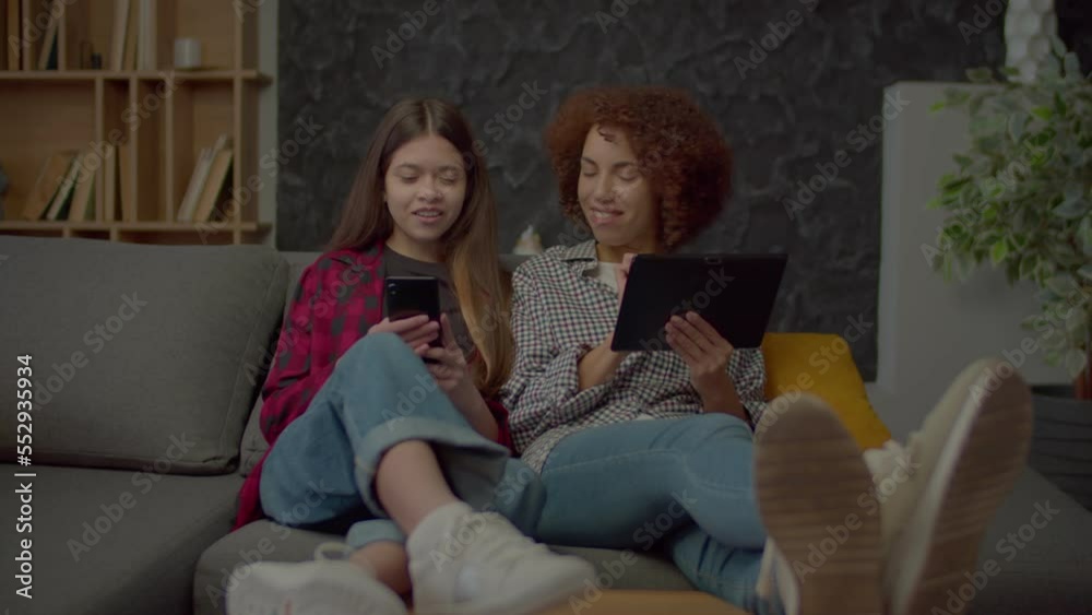 Positive cute multicultural adolescent daughter and attractive African American mother relaxing on sofa, networking and browsing social media content online using smart technology devices indoors. Positive cute multicultural adolescent daughter and attractive African American mother relaxing on sofa, networking and browsing social media content online using smart technology devices indoors.