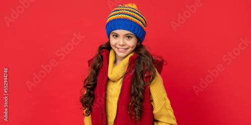 glad kid in knitted winter hat and down vest on red background, childhood