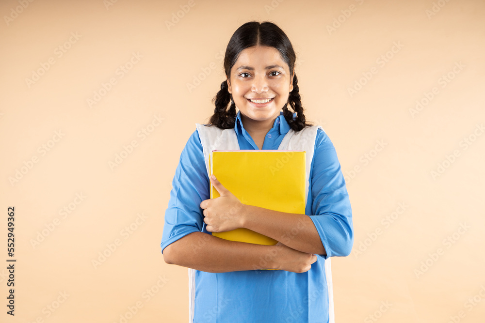 Happy Indian student girl wearing blue school uniform holding books standing isolated over beige background, Education and knowledge concept. Happy Indian student girl wearing blue school uniform holding books standing isolated over beige background, Education and knowledge concept.