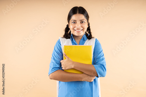 Happy Rural Indian student schoolgirl wearing blue government school uniform holding books and bag standing isolated over beige background, Studio shot,closeup, Education concept.