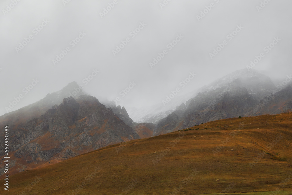 Atmospheric misty landscape with fuzzy silhouettes of sharp rocks in low clouds during rain. Dramatic view to large mountains blurred in rain haze in gray low clouds.