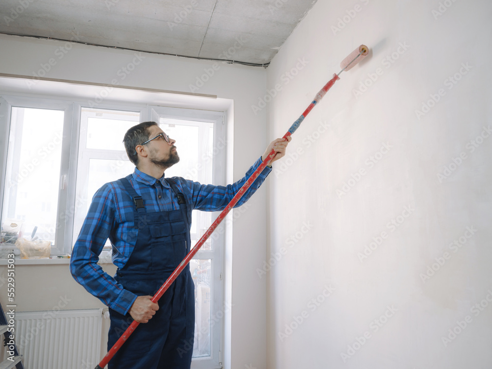 young man in blue work suit hold paint roller, over white wall room background. Instruments accessories for renovation apartment room. Repair home concept young man in blue work suit hold paint roller, over white wall room background. Instruments accessories for renovation apartment room. Repair home concept