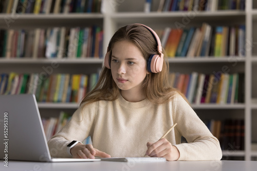 Serious hardworking smart student girl in wireless headphones working on creative essay, project, study research in college public library, watching online video lesson on internet, writing notes