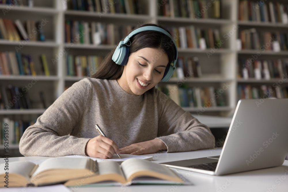 Happy Latin student girl in wireless earphones studying in public library, writing notes at laptop, open books, listening music for focus, watching learning class, webinar, lesson, making summary Happy Latin student girl in wireless earphones studying in public library, writing notes at laptop, open books, listening music for focus, watching learning class, webinar, lesson, making summary