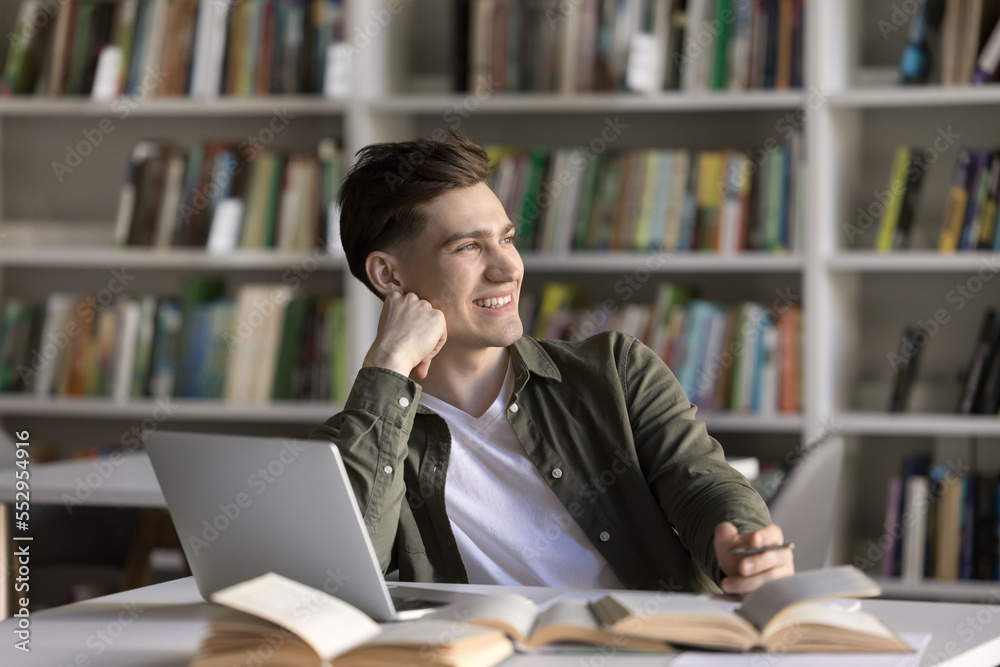 Happy dreamy student guy working on scientific article, study research in library, writing notes at laptop, looking away in deep thoughts, thinking over creative ideas for essay, smiling Happy dreamy student guy working on scientific article, study research in library, writing notes at laptop, looking away in deep thoughts, thinking over creative ideas for essay, smiling