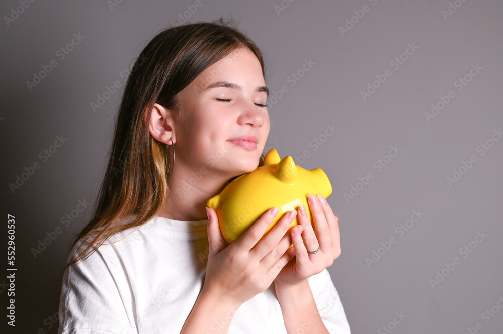 Cute teenage girl smiling while holding and hugging a piggy bank. Close up portrait on gray background Cute teenage girl smiling while holding and hugging a piggy bank. Close up portrait on gray background