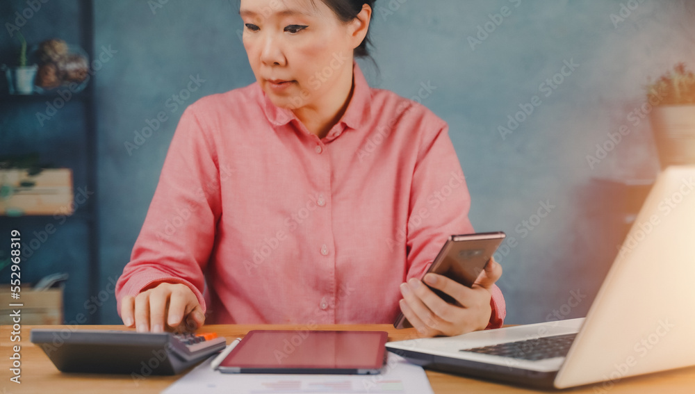 .Portrait of Asian young woman use smartphone working with laptop at Home, checking bills and calculating sum of month home expenses, paying bills online Startup business financial calculate account.. .Portrait of Asian young woman use smartphone working with laptop at Home, checking bills and calculating sum of month home expenses, paying bills online Startup business financial calculate account..