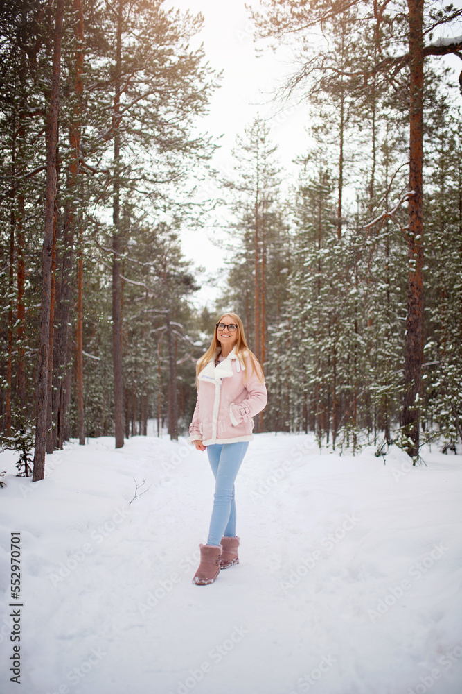 Happy young blonde woman in glasses, pink jacket in winter clothes on background of snowy forest
