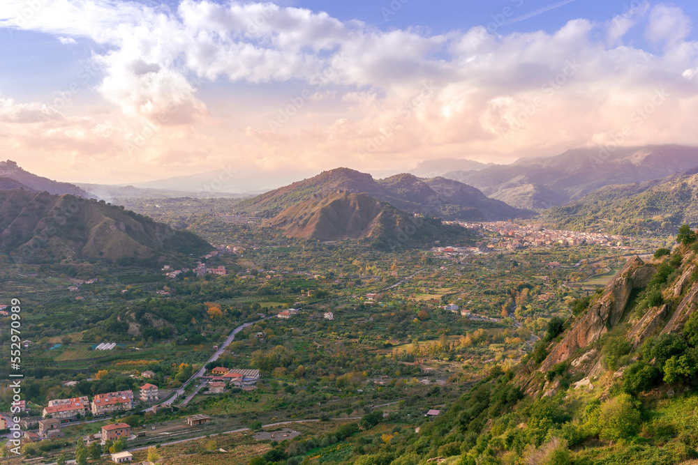 Fototapeta premium beautiful landscape with amazing view from highland with green slopes and bushes to a valley with town, road and nice mountains and scenic cloudy sunset on background