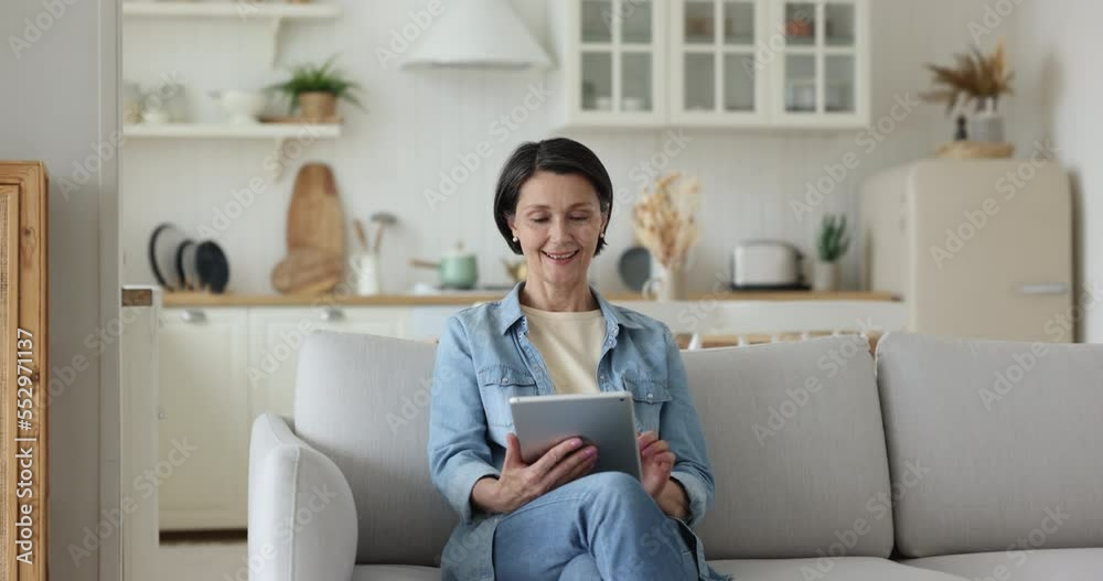 Smiling attractive older female sit on cozy sofa using digital tablet, buying goods and gifts on-line, makes easy comfort purchase distantly through web retail electronic services. E-commerce, tech Smiling attractive older female sit on cozy sofa using digital tablet, buying goods and gifts on-line, makes easy comfort purchase distantly through web retail electronic services. E-commerce, tech