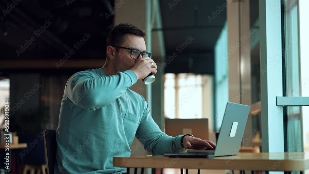 Man working on laptop and drinking coffee from a paper cup. Positive smiling businessman typing on computer sitting in comfortable office. Man working on laptop and drinking coffee from a paper cup. Positive smiling businessman typing on computer sitting in comfortable office.