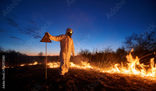 Fototapeta Naklejka Na Ścianę i Meble -  Fireman ecologist fighting fire in field at night. Man in protective suit and gas mask near burning grass with smoke, holding warning sign with skull and crossbones. Natural disaster concept.