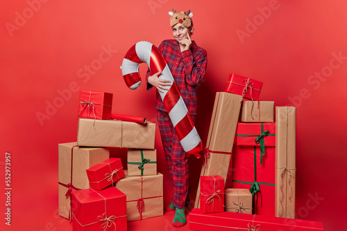 Indoor shot of beautiful European woman wears reindeer sleepmask and comfortable pajama poses with inflated candy cane stands around wrapped gift boxes against bright red background. Winter holidays photo