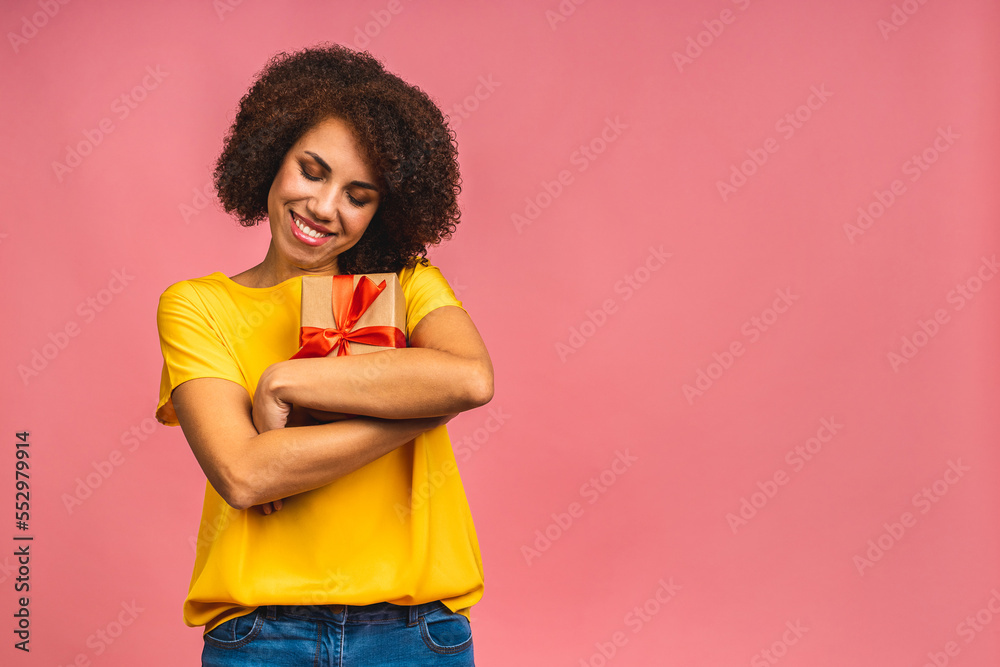Happy young beautiful african american lady in casual laughing while holding present gift box isolated over pink background.