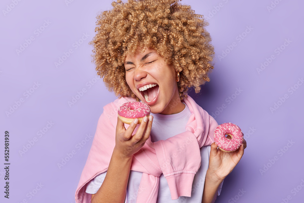 Harmful nutrition unhealthy sweet food concept. Curly haired woman eats delicious doughnuts with appettite enjoys favorite dessert keeps mouth widely opened dressed casually isolated over purple wall Harmful nutrition unhealthy sweet food concept. Curly haired woman eats delicious doughnuts with appettite enjoys favorite dessert keeps mouth widely opened dressed casually isolated over purple wall