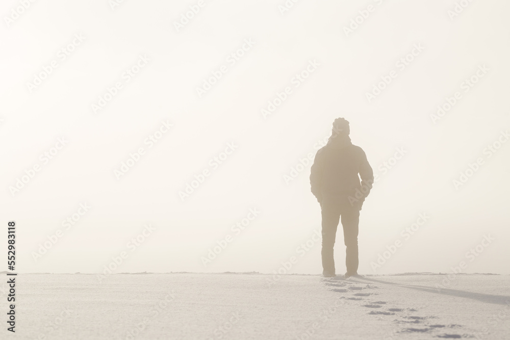 Young adult man silhouette standing in nature mist and looking far away. Thinking about life. Back view. Fresh human boots footprints in fresh deep snow. Cold snowy winter day. Empty place for text. - obrazy, fototapety, plakaty 