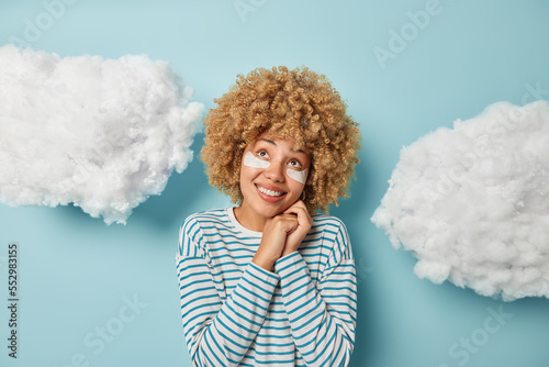 Indoor shot of curly haired woman keeps hands near face smiles toothily focused above dreams about something wears casual striped jumper applies beauty patches under eyes to reduce wrinkles.