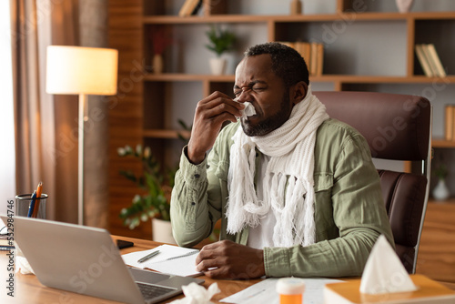 Unhappy sad adult black man with scarf blows his nose in napkin, suffers from runny nose and fever, works with computer