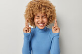 Positive curly haired woman crosses fingers for good luck keeps eyes closed smiles broadly believes dreams come true wears blue turtleneck anticipates for something isolated over grey background