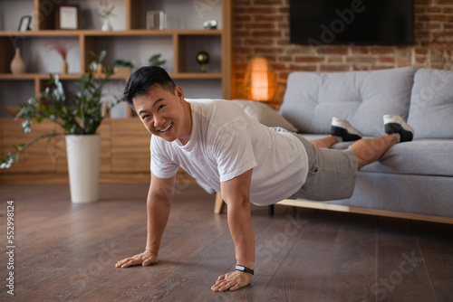Strong middle aged asian man doing push ups with legs on sofa, working out at home and smiling at camera © Prostock-studio