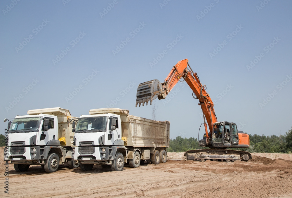 orange crawler excavator and two gray construction dump trucks in the process of loading and transporting soil in a quarry against a blue sky. Commercial vehicles. Construction site, earthworks