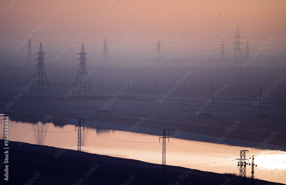 High voltage power lines and transmission towers by the river at sunset ...