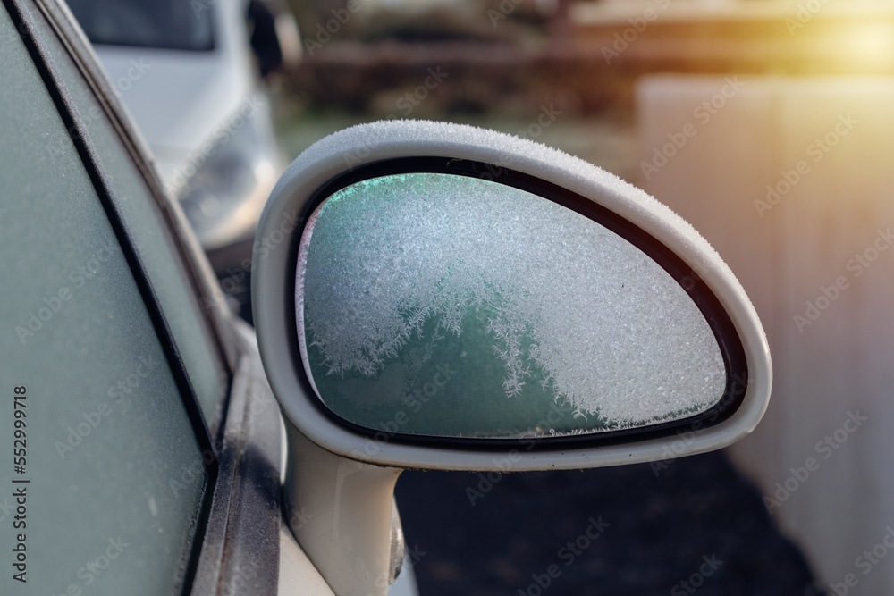Frozen white rearview mirror of a parked car in a outdoor parking, early winter frost
