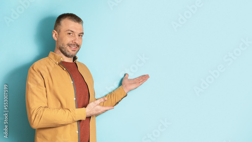 Happy mature caucasian man showing blank space aside, gesturing with hands and smiling, standing over blue background