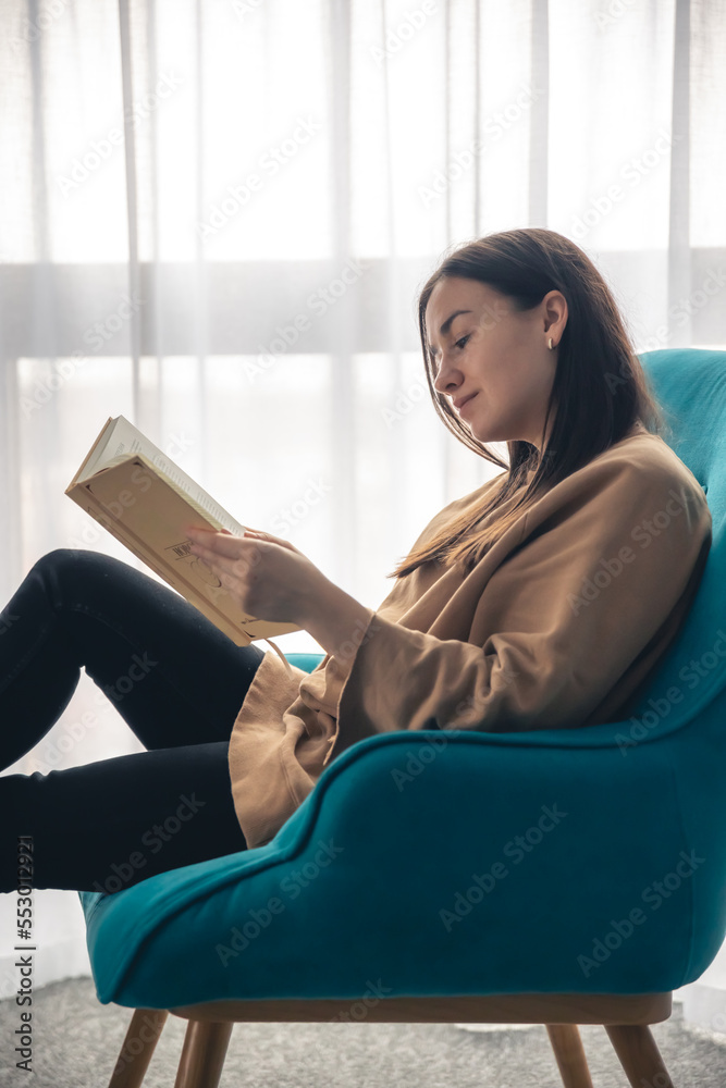 A young woman is reading a book while sitting on an armchair by the window. A young woman is reading a book while sitting on an armchair by the window.