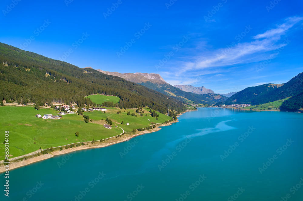 Fototapeta premium Aerial view of lake (Reschensee). Large reservoir surrounded by mountains at sunny noon. Recreation area for tourists and sportsmen. Organic farm on the shore. Italy, Vinschgau, Giern.