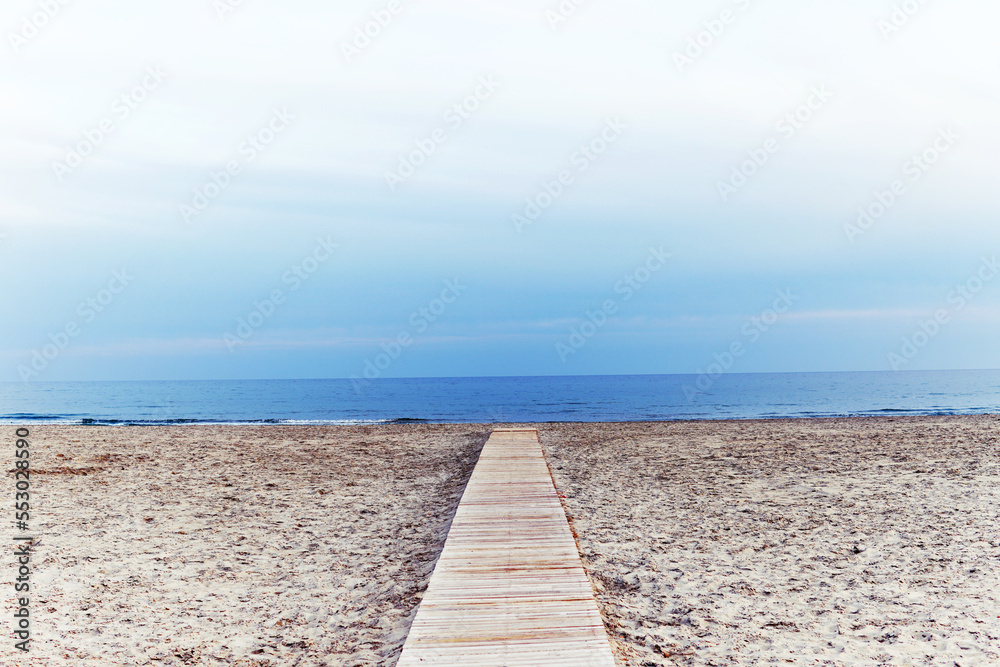 Empty sandy beach with blue sky in cold weather. Wooden path in the middle leading to the blue ocean sea.