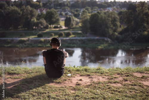 A young man in headphones sits on the river bank in the park, listens to music from a smartphone and contemplates nature.
