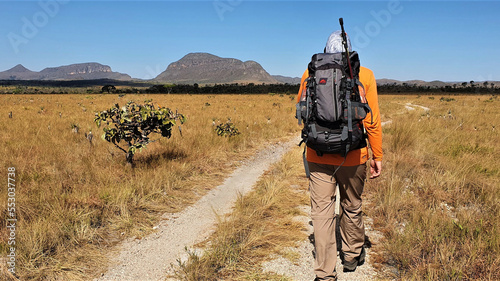 man with backpack on his shoulders walking along a trail in the savannah with mountains in the background and blue sky