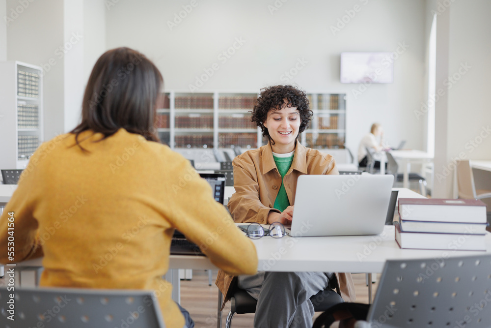 Fototapeta premium two women in library or coworking are sitting opposite each other working on laptop. meeting and negotiations of two colleagues, classmates. scientific work in office or university. success, career