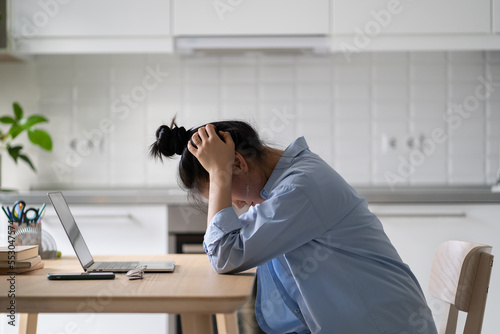 Stressed frustrated female freelancer sitting in front of laptop in kitchen, holding head in hands, struggling with task while working remotely. Work from home burnout, emotional side of freelancing