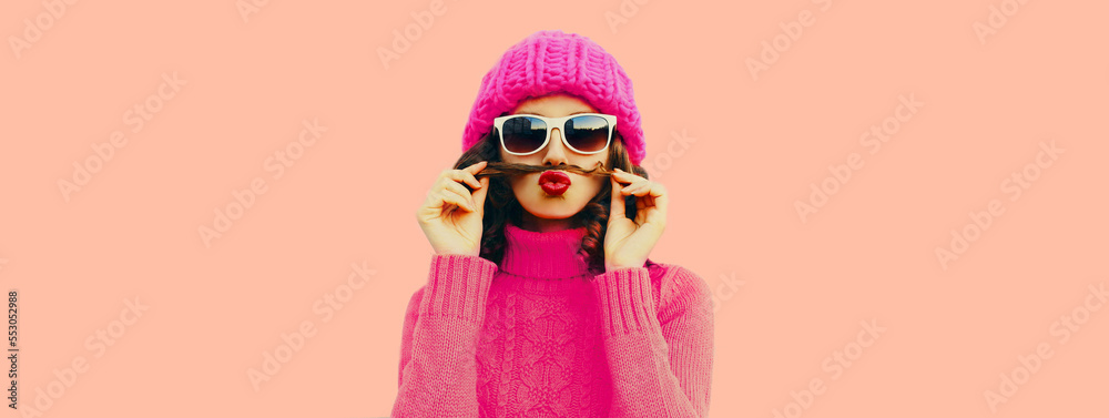 Portrait of funny young woman showing mustache with hair blowing her red lips having fun wearing knitted pink hat, sweater on pink background Portrait of funny young woman showing mustache with hair blowing her red lips having fun wearing knitted pink hat, sweater on pink background
