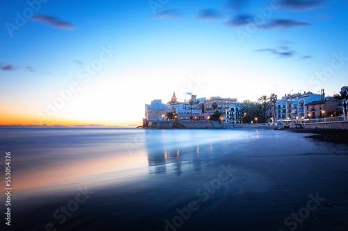 Fotografia de larga exposición de Sitges desde la playa San Sebastià con la iglésia de Sant bartomeu iluminada al fondo y un cielo con nubes al atardecer photo