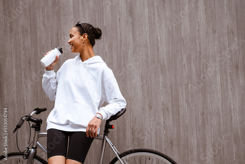 Positive african american woman in hoodie and cycling shorts holding sports bottle near bike on street 