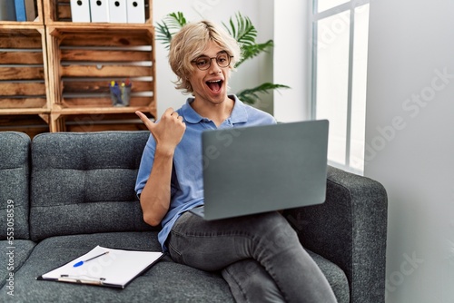 Young man working using computer laptop sitting on the sofa pointing thumb up to the side smiling happy with open mouth