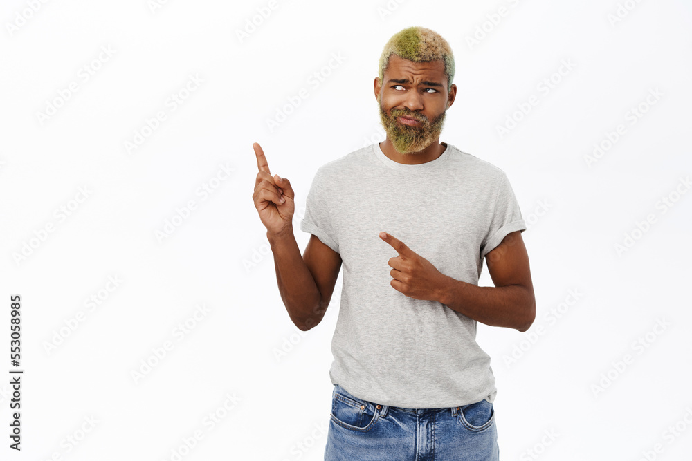 Naklejka premium Upset hipster guy with yellow beard and hair, points left, frowns and looks sad, stands over white background