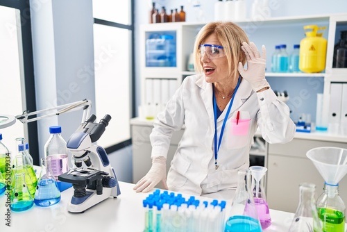 Middle age blonde woman working at scientist laboratory smiling with hand over ear listening an hearing to rumor or gossip. deafness concept.