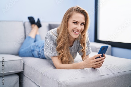 Young blonde woman using smartphone lying on sofa at home