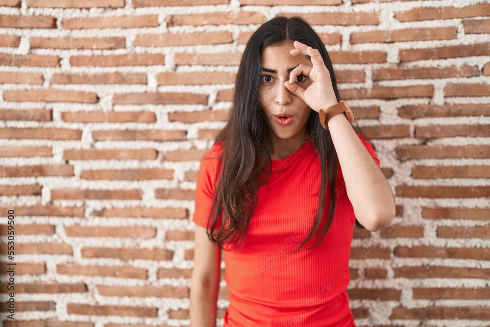 Young teenager girl standing over bricks wall doing ok gesture shocked with surprised face, eye looking through fingers. unbelieving expression.